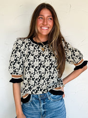 Woman wearing a black and white floral blouse with ruffled sleeves and brown stripe detail on sleeves and bottom against a white wall.