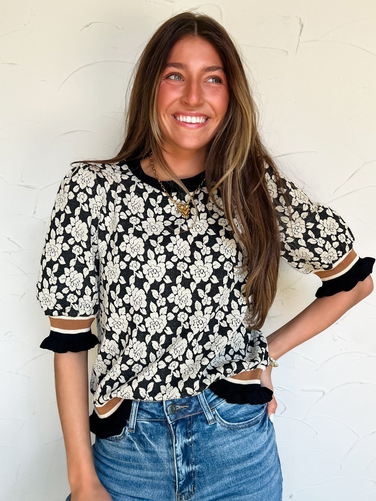 Woman wearing a black and white floral blouse with ruffled sleeves and brown stripe detail on sleeves and bottom against a white wall.