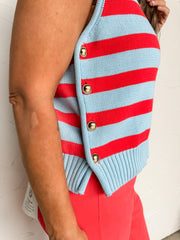 A woman smiling against a textured off-white wall, wearing a sleeveless sweater vest crafted from a 100% cotton knit construction in a light blue and bright red striped pattern. The top features a round crew neckline, a classic horizontally striped body, and a ribbed hemline. A distinctive side-button panel closure along the lower right hem is detailed with four polished, dome-shaped gold buttons. She is styled with a cream trucker hat that reads "America's Sweetheart", layered gold necklaces, and coordinat