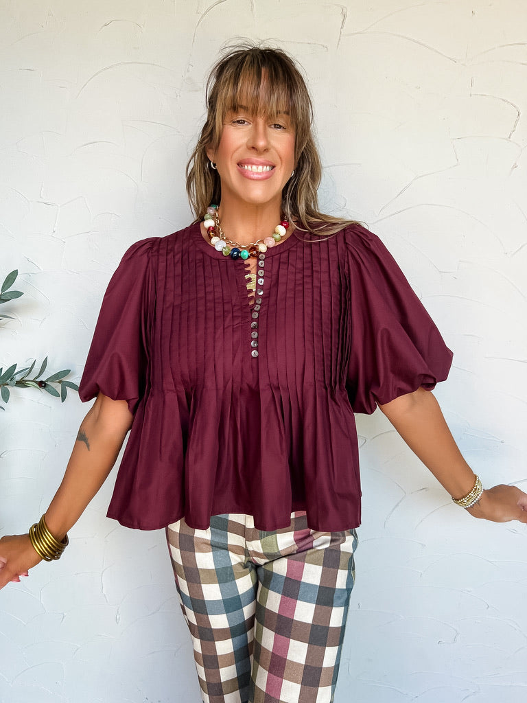 A three-quarter length photograph of a smiling woman with long brown hair and bangs posing with her arms slightly outstretched against a textured off-white wall. She is wearing a short-sleeved deep burgundy blouse made from an 80% polyester and 20% cotton blend, featuring vertical pintuck pleat detailing on the front bodice, a small buttoned split placket neckline, dramatic volume puff sleeves, and a flared hemline, paired with colorful plaid check trousers in shades of blue, burgundy, brown, and cream, sty