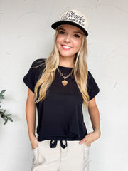 A medium shot of a smiling blonde woman standing against a textured white wall. She is wearing a basic black crew-neck t-shirt featuring rolled, cuffed short sleeves and a casual drawstring tie design at the center hem. She has her hands in the pockets of light beige linen trousers and is accessorized with a gold chain heart necklace and a cream and black trucker hat that reads "Blondes HAVE MORE FUN".