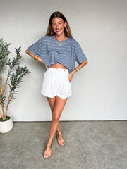 Woman wearing a blue striped shirt and white shorts standing against a white wall with a plant to her left.