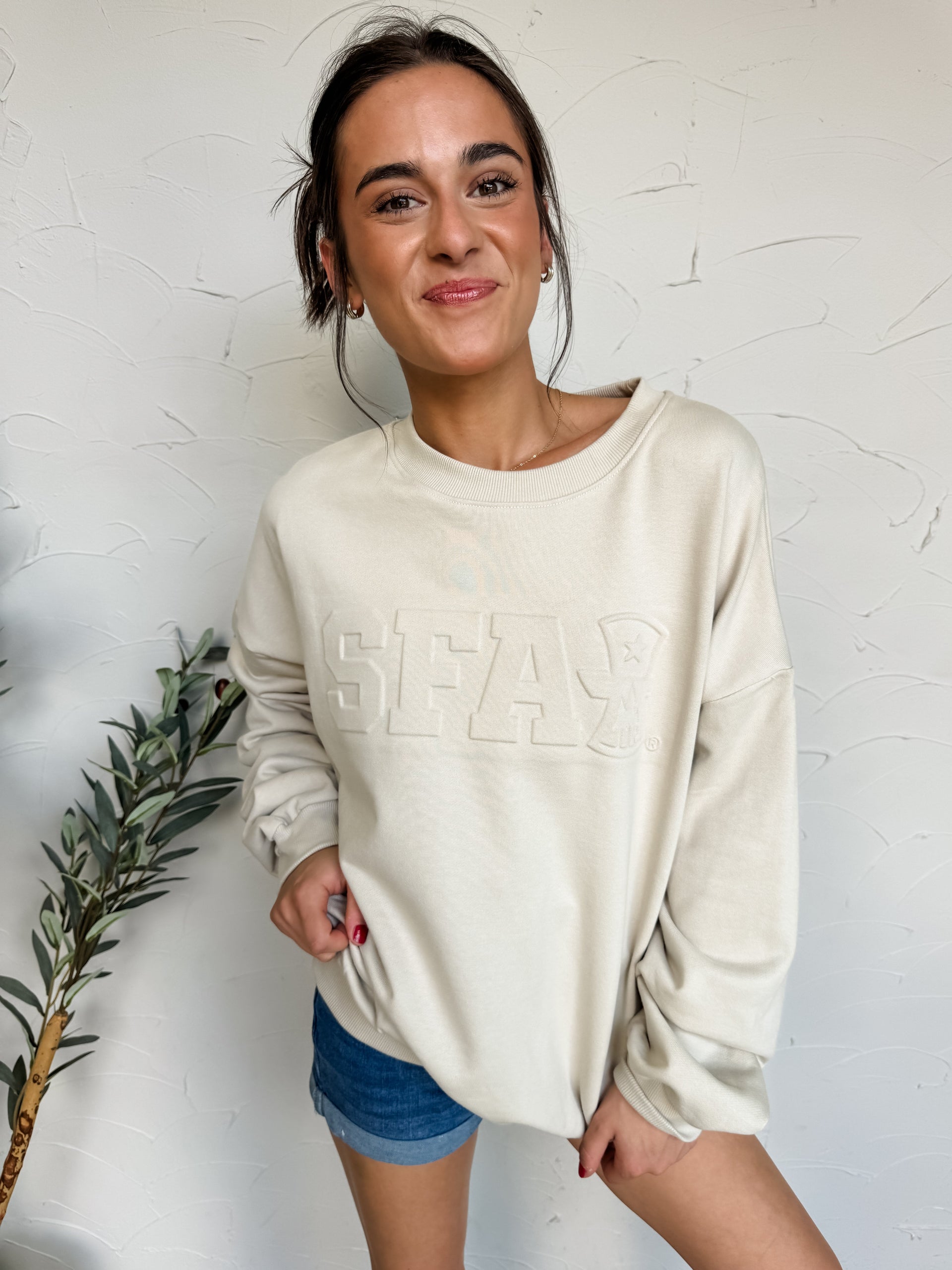 Woman wearing a beige sweatshirt with 'SFA' text against a white wall.