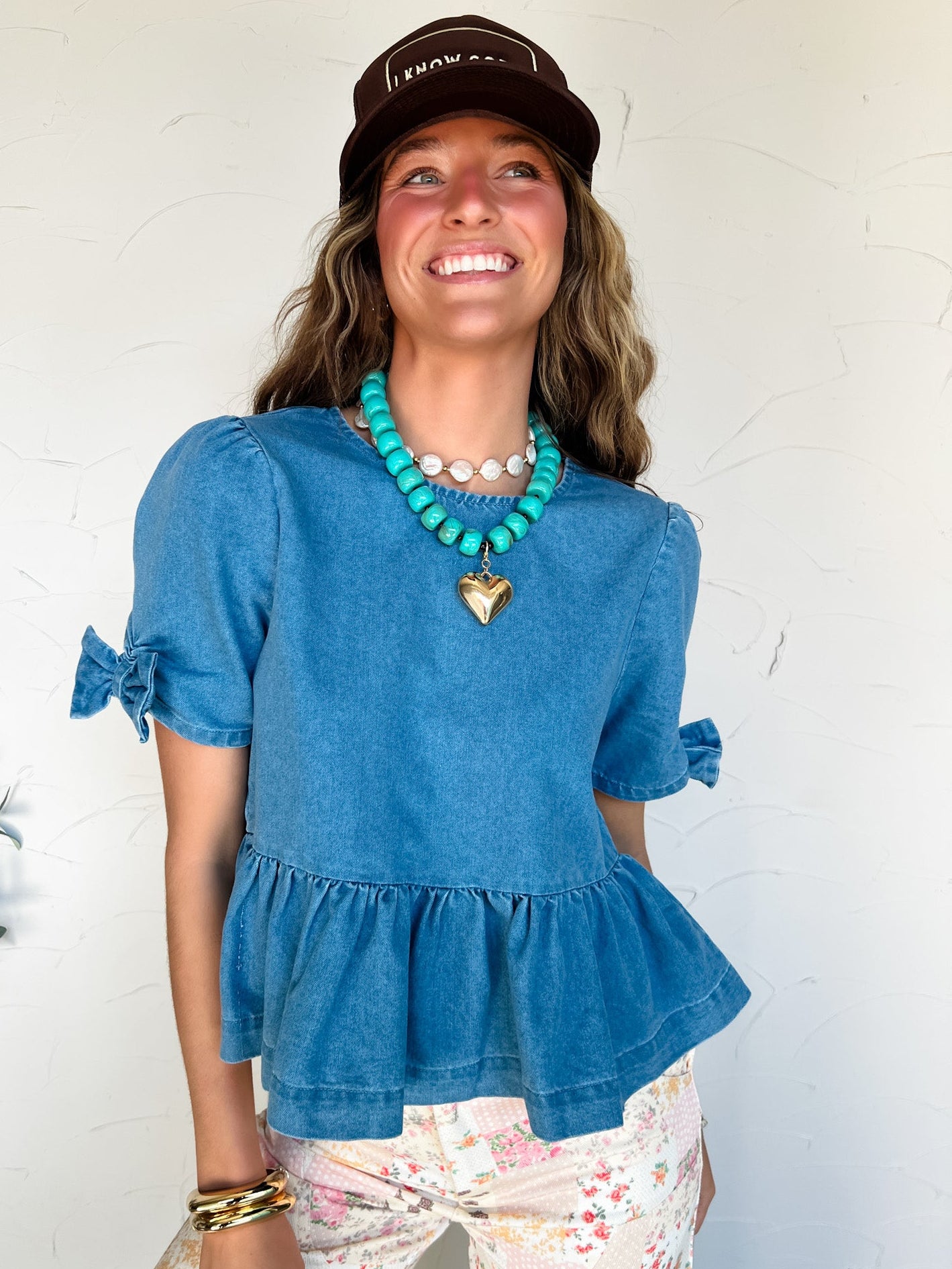 Woman wearing a denim blue peplum top and floral pants with a necklace, standing against a white wall.