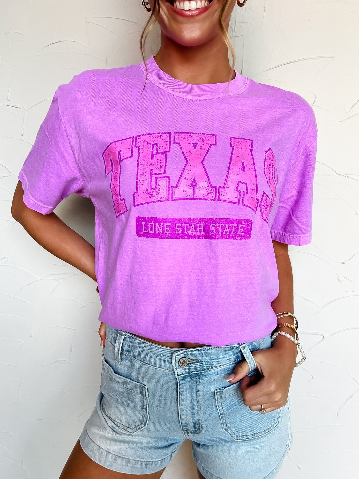 Person wearing a purple 'TEXAS Lone Star State' t-shirt and denim shorts on a white background