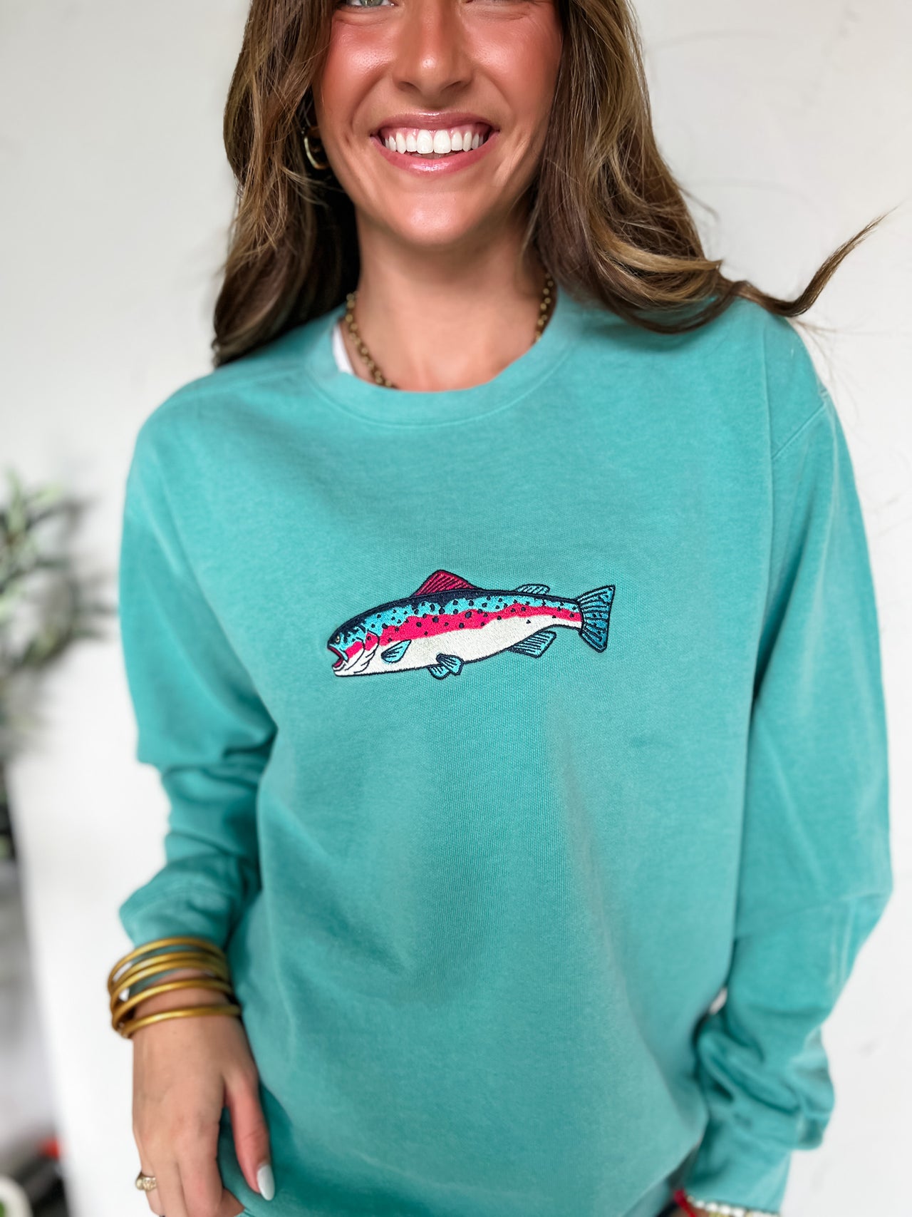 Woman wearing a teal sweatshirt with a embroidered fish design, smiling against a white background.