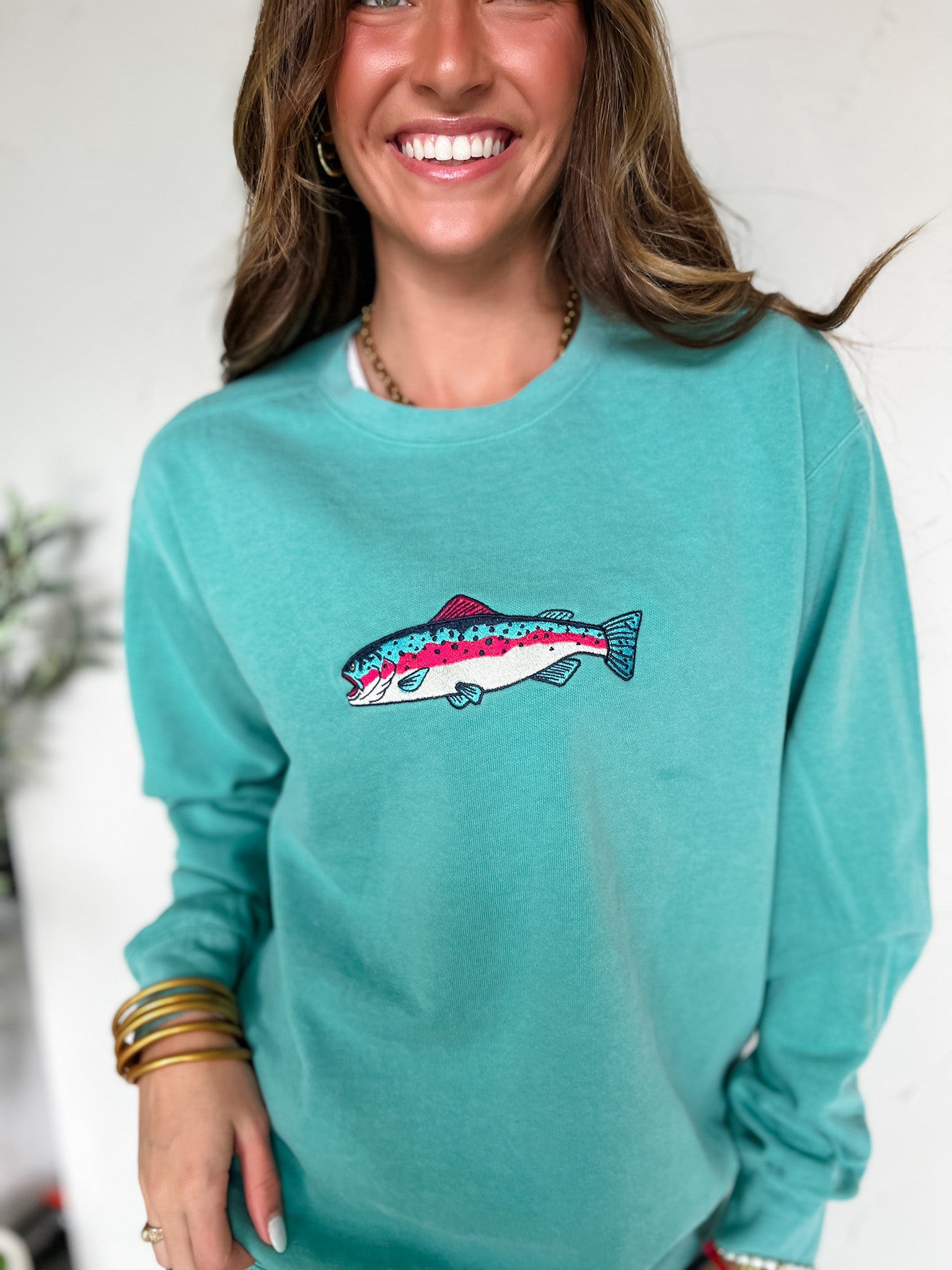 Woman wearing a teal sweatshirt with a embroidered fish design, smiling against a white background.
