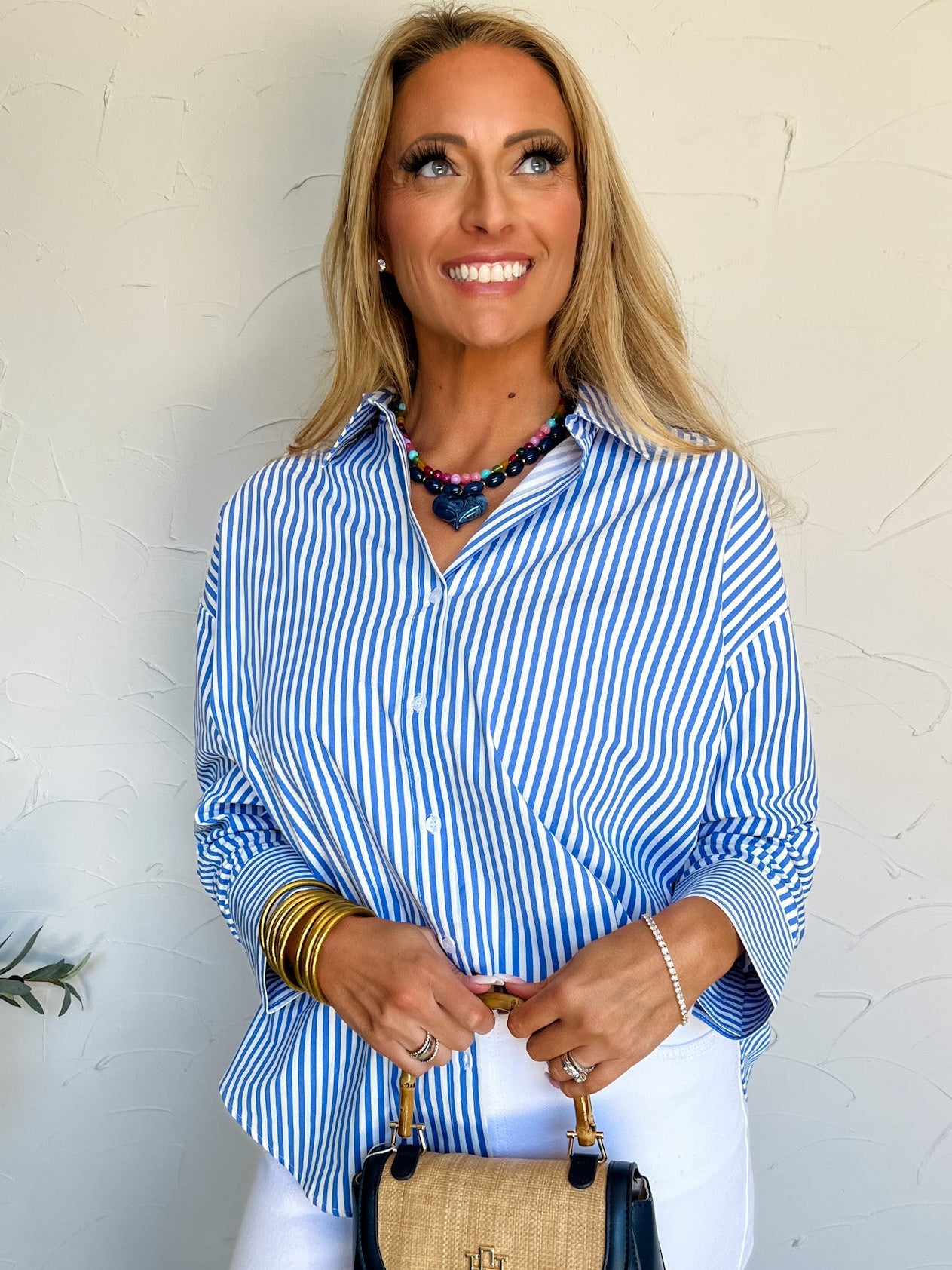 Woman wearing a blue and white striped shirt with a black and straw handbag against a white wall.
