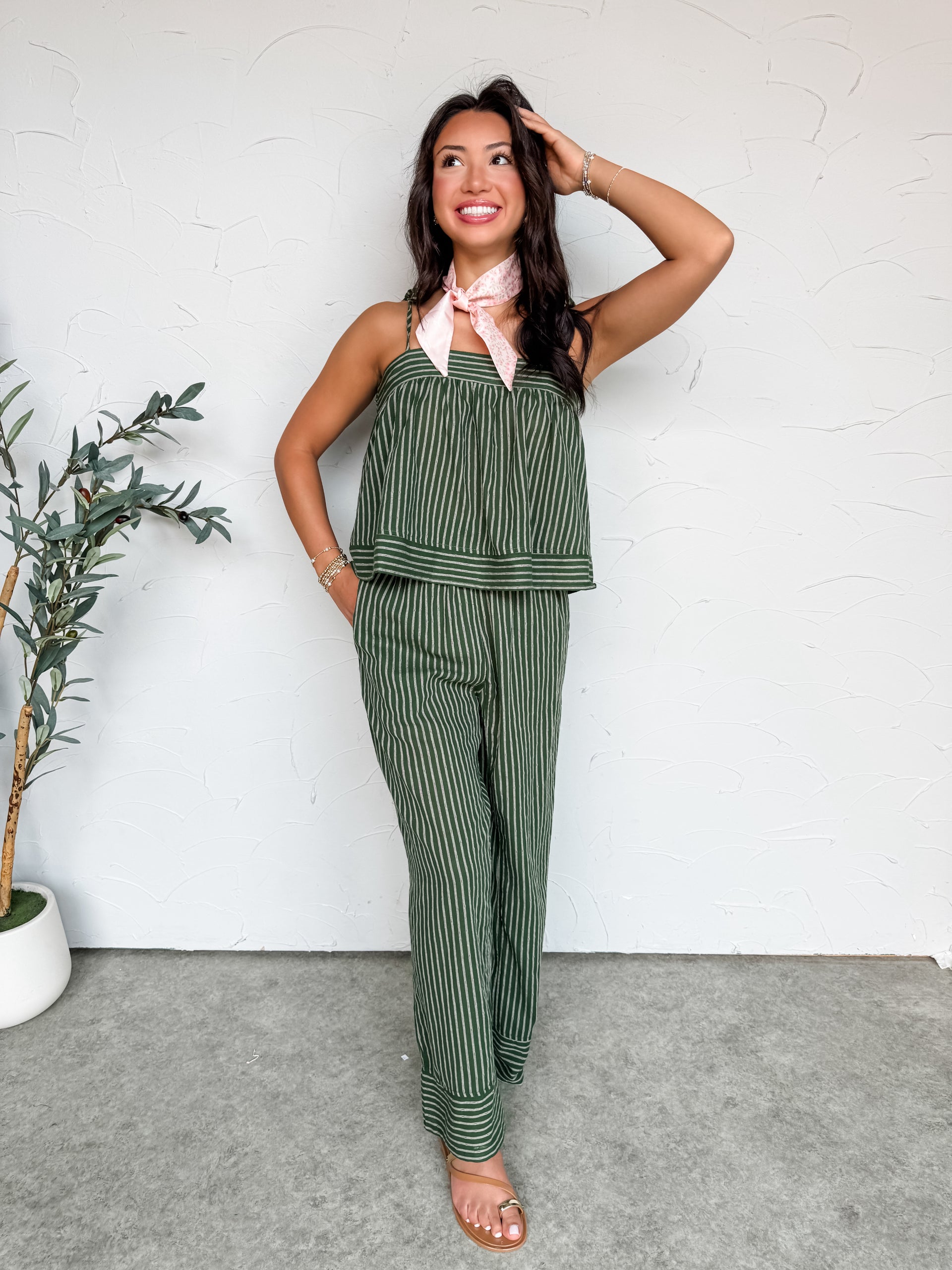 A full-length photo of a smiling woman standing against a textured white wall, showcasing a matching two-piece olive green and white pinstriped lounge set. She is playfully posing with one leg lifted, revealing a loose, wide-leg trouser fit. The top is a flowy camisole with self-tie shoulder straps, styled with a light pink neck scarf. A potted olive plant sits to her left, and she completes the look with simple flat sandals.