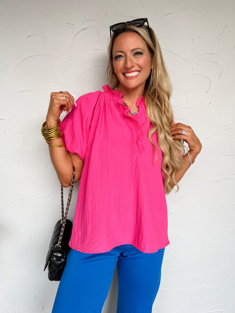 Woman wearing a bright pink top and blue pants against a white wall.