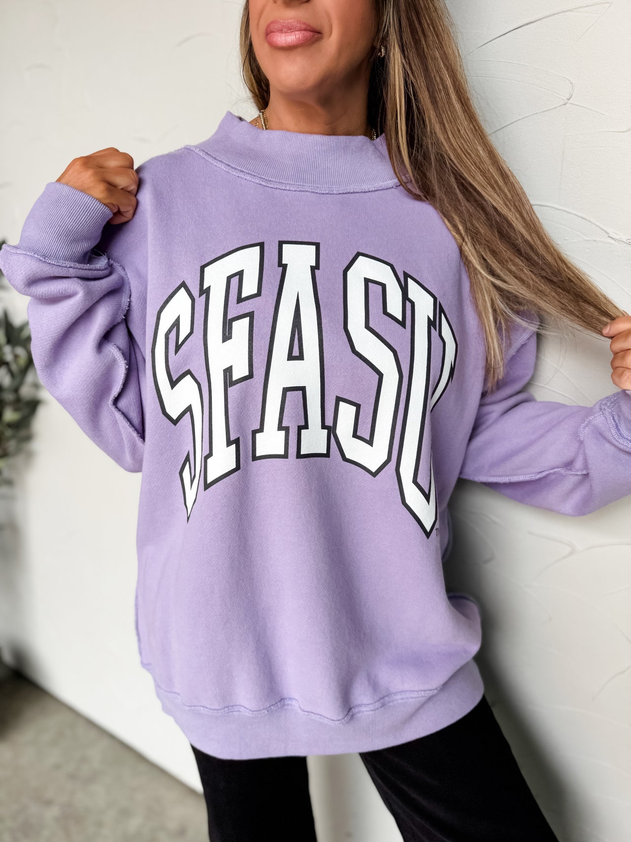 Person wearing a purple sweatshirt with 'SEAS' text against a white wall.