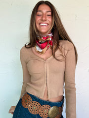 Woman wearing a beige ribbed sweater, blue skirt with a large belt, and a colorful scarf against a white wall.