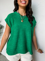 A close-up photograph from the chin down of a smiling brunette woman posing against a textured white wall. She is wearing an oversized crewneck knit top made from a textured 75% acrylic and 25% nylon blend sweater fabric in a vibrant green colorway, featuring a garter stitch pattern, sleeveless cap shoulders, and small side hem slits.