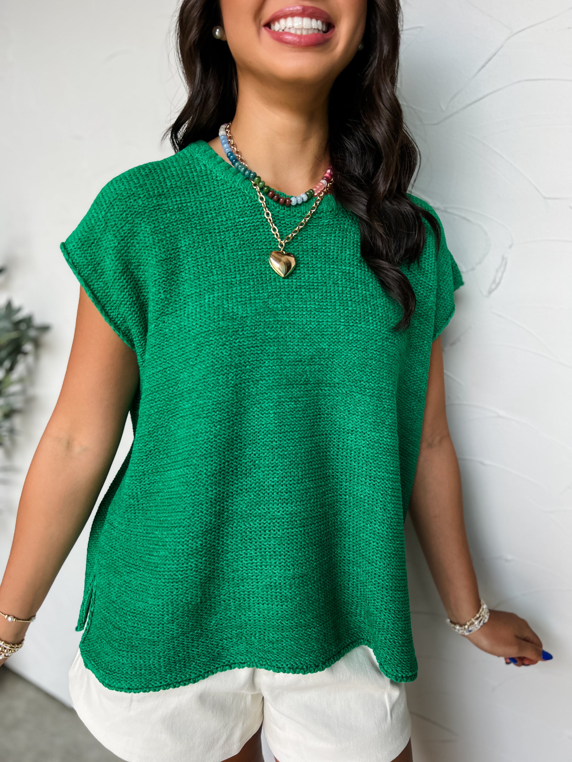 A close-up photograph from the chin down of a smiling brunette woman posing against a textured white wall. She is wearing an oversized crewneck knit top made from a textured 75% acrylic and 25% nylon blend sweater fabric in a vibrant green colorway, featuring a garter stitch pattern, sleeveless cap shoulders, and small side hem slits.
