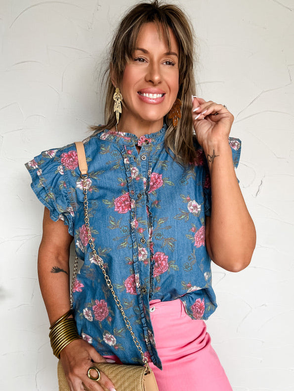 Woman wearing a blue floral top and pink pants, holding a beige handbag against a white background.