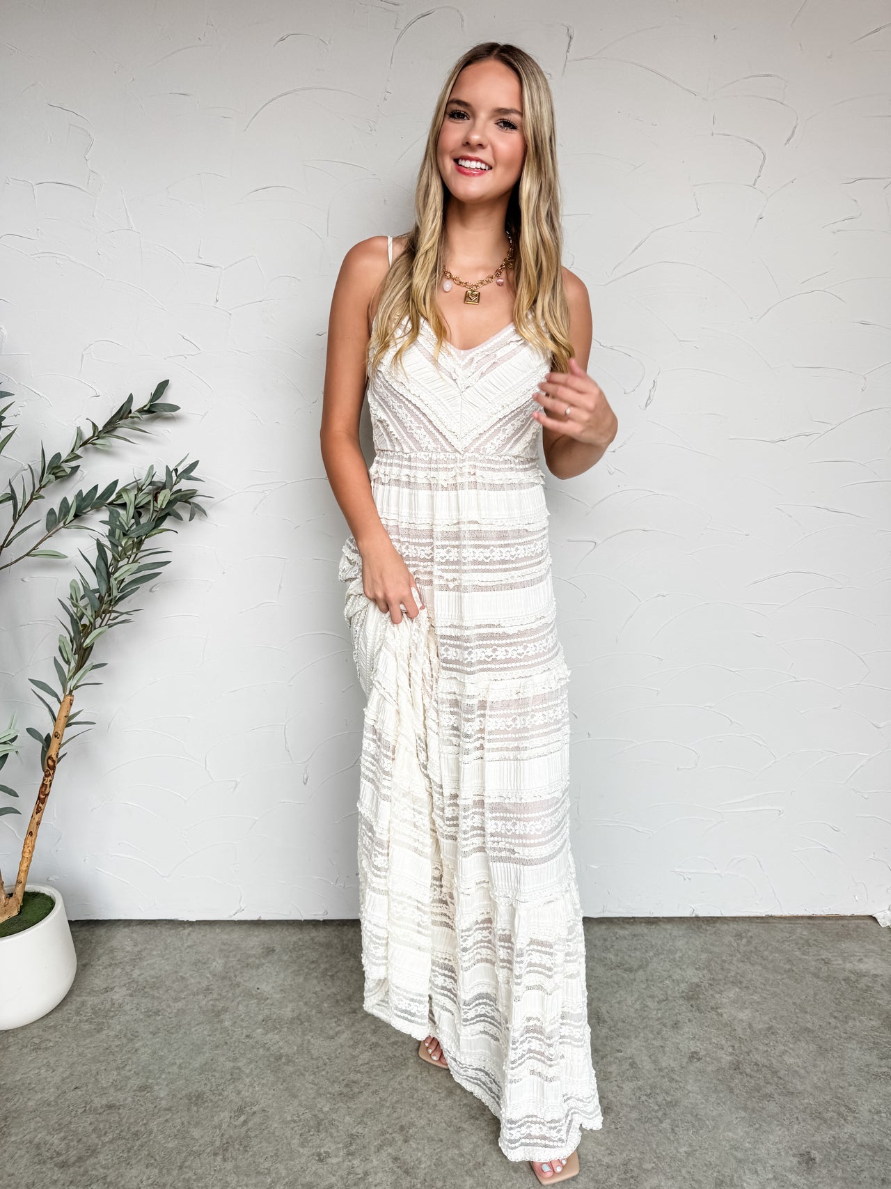 A smiling blonde woman stands against an off-white textured wall, wearing a cream-colored lace maxi dress. The floor-length dress features a V-neckline with delicate spaghetti straps, geometric chevron patterned lace on the bodice, and a tiered skirt with alternating rows of sheer patterned lace and solid micro-pleated ruffles. She is styled with a chunky gold link necklace with a square pendant and simple flat sandals.