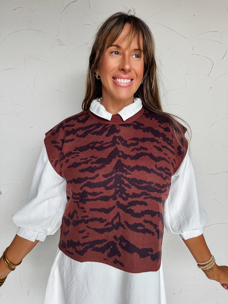 A three-quarter length photograph of a smiling woman with long brown hair with bangs posing against a textured white wall. She is wearing a fine-knit sleeveless sweater vest featuring a dark rust brown and black tiger stripe animal print made from a 50% viscose, 30% polyester, and 20% nylon blend, layered over a crisp white collared mini dress with puff sleeves, styled with stacked gold bangles and delicate beaded bracelets.