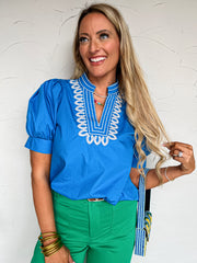 Woman wearing a blue blouse with white embroidery and green pants against a white wall.