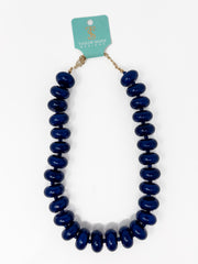 A chunky beaded necklace with glossy dark blue oval beads, attached to a light teal paper tag that reads 'Taylor Shave Designs,' shown on a plain white background.