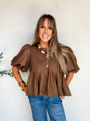 A portrait photograph of a smiling woman with long brown hair and bangs posing with her hands in her pockets against a textured off-white wall next to a potted olive plant. She is wearing a short-sleeved chocolate brown blouse made from an 80% polyester and 20% cotton blend, featuring vertical pintuck pleating across the chest, a partial button front, dramatic puff sleeves, and a flared peplum hem, paired with medium wash jeans, styled with a colorful beaded strand necklace, a gold pendant, and stacked gold