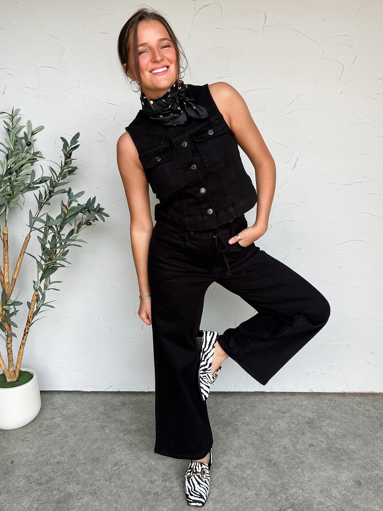 Woman in black sleeveless top and pants posing against a white wall with a plant to her left.