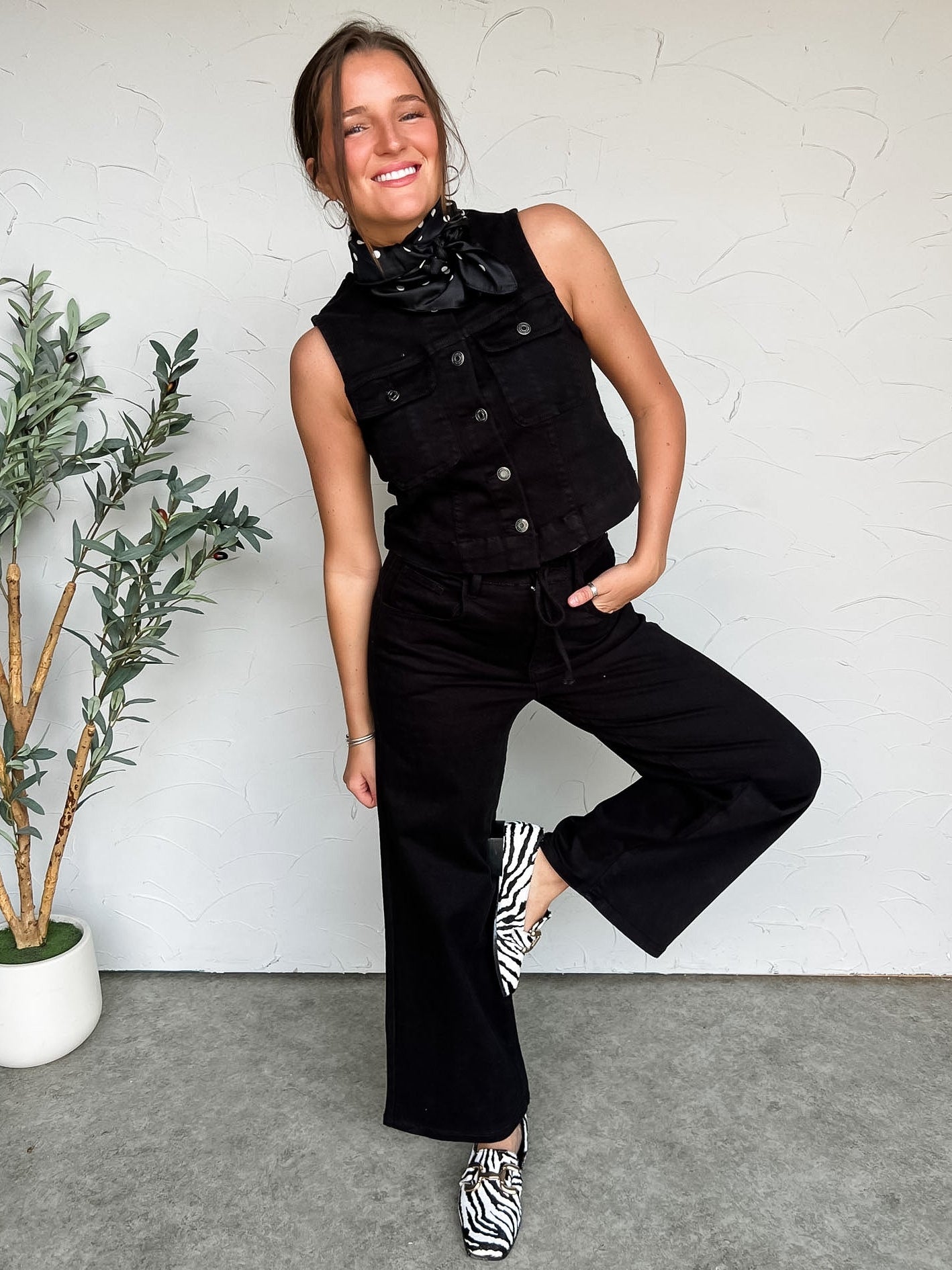 Woman in black sleeveless top and pants posing against a white wall with a plant to her left.