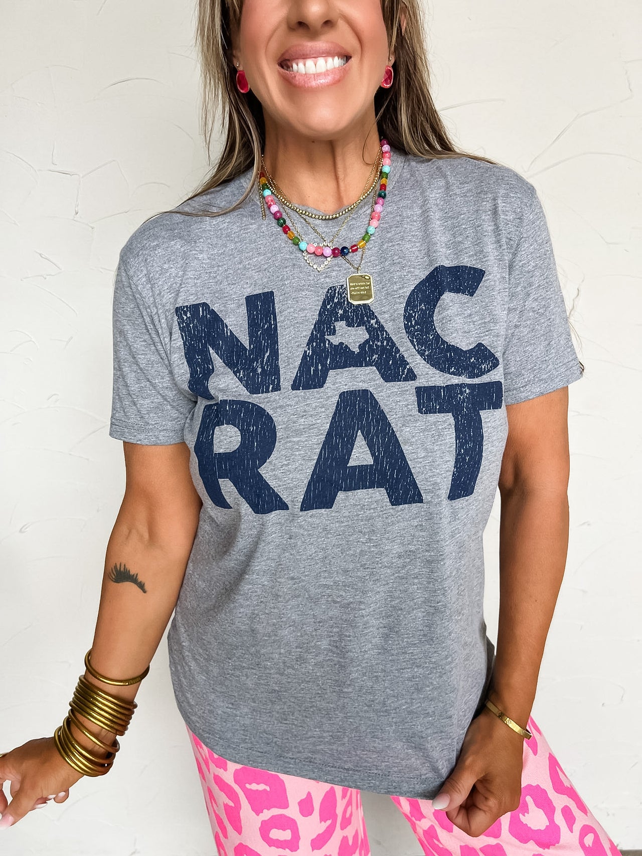Person wearing a gray t-shirt with 'NAC RAT' text, standing against a white wall.