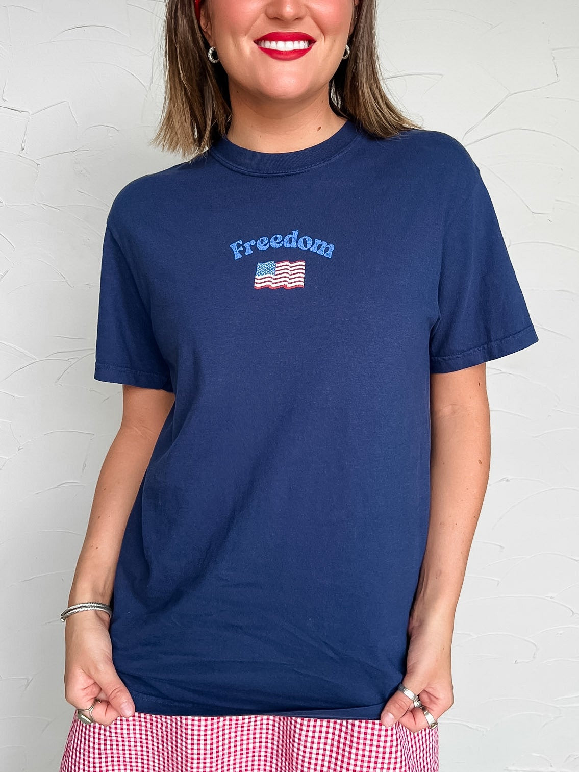 Woman wearing a navy blue t-shirt with 'Freedom' and an American flag design, standing against a white wall.