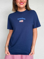 Woman wearing a navy blue t-shirt with 'Freedom' and an American flag design, standing against a white wall.