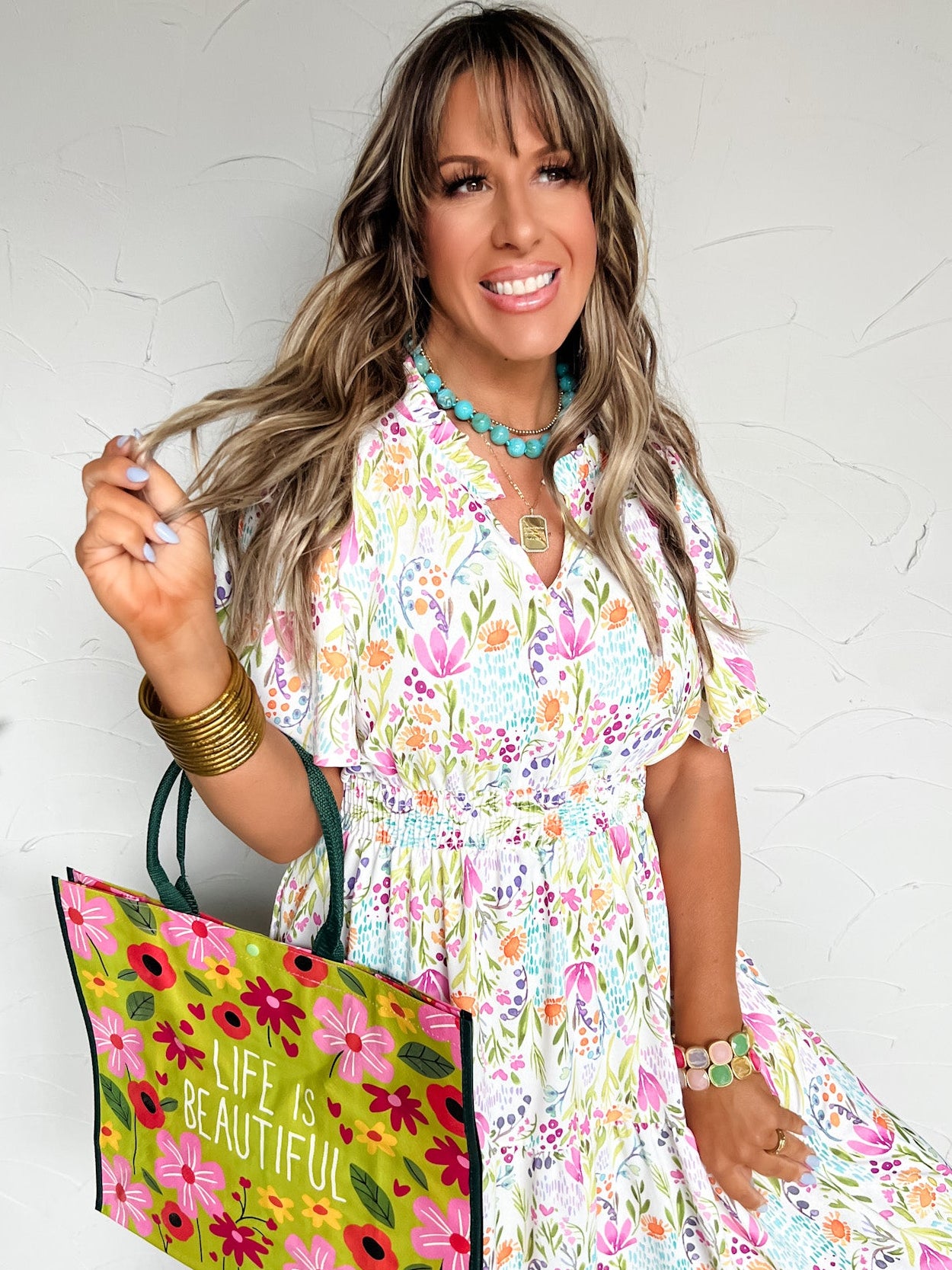 Woman in a floral dress holding a colorful tote bag with 'Life is Beautiful' text against a white wall.