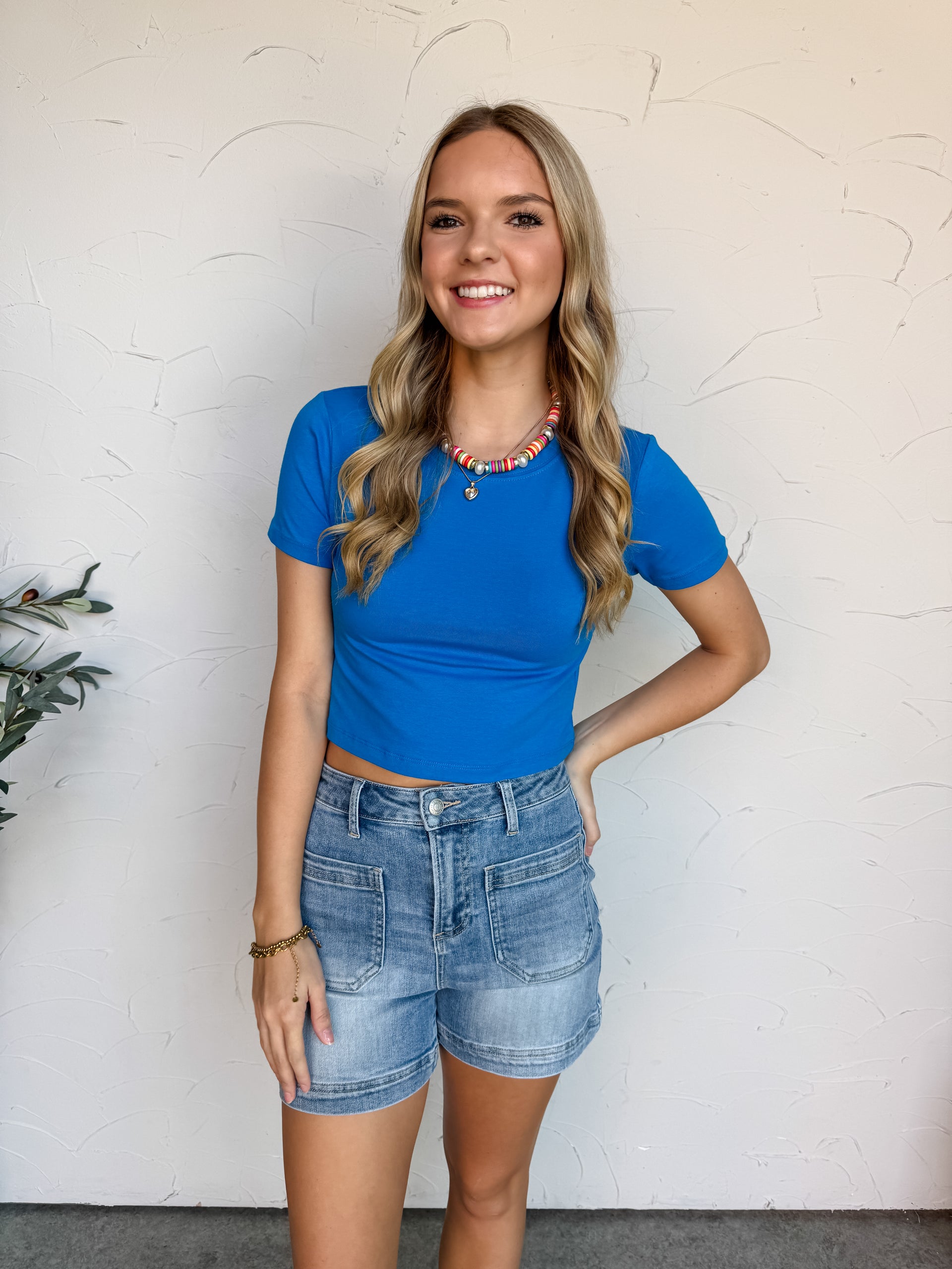 Woman wearing a blue top and denim shorts against a white wall