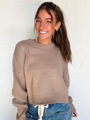 Woman wearing a brown sweater and blue jeans against a white wall