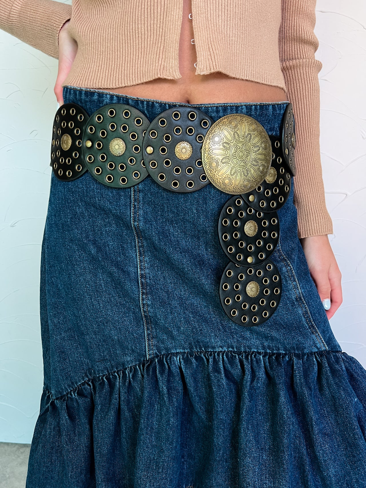 Model wearing a Western-inspired belt with faux-leather discs and an antique gold center over a denim skirt.