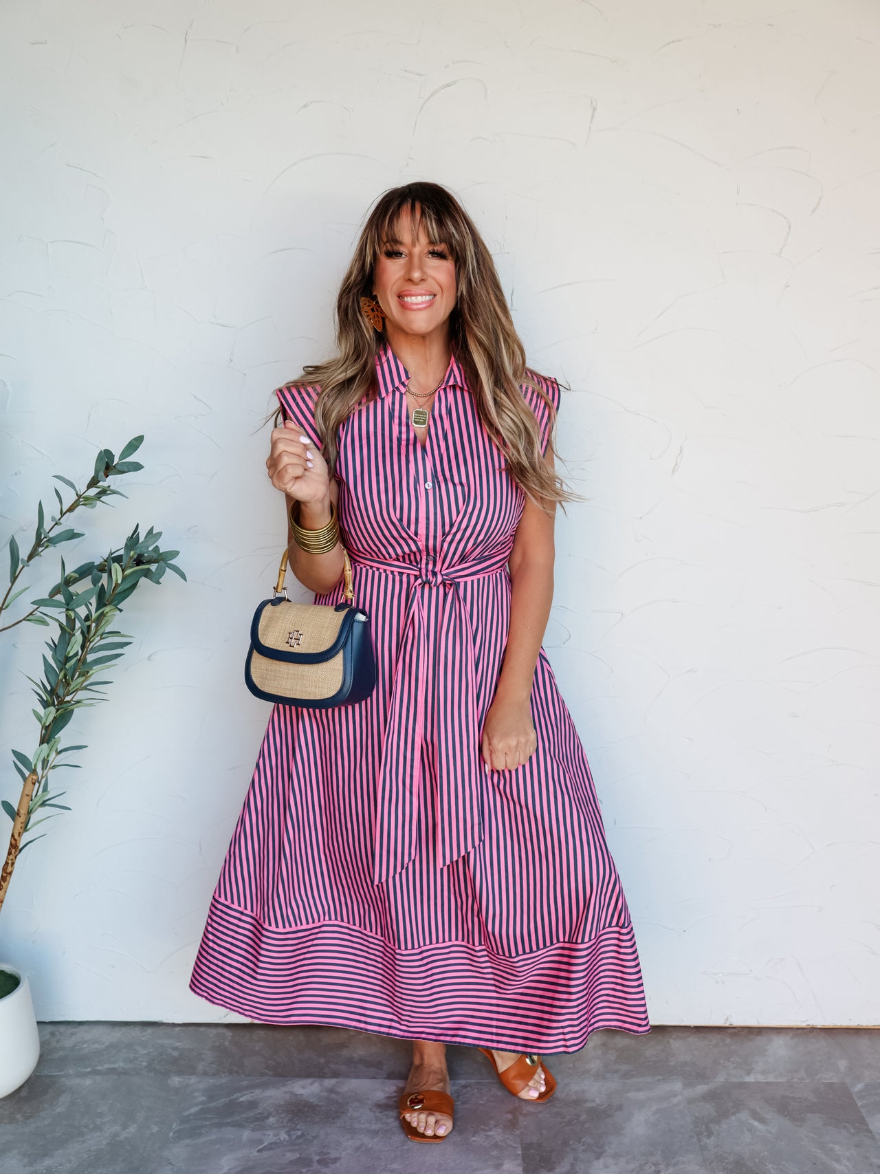 A full-length photograph of a smiling brunette woman with bangs posing against a textured white wall. She is wearing a button-up dark pink and navy blue vertically striped midi shirt dress made from a 100% cotton fabric, featuring a collar, cap sleeves, a matching tie belt at the waist, and horizontal stripes at the hemline, styled with brown slides and a navy and straw handbag.