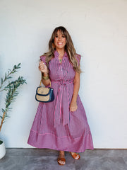 A full-length photograph of a smiling brunette woman with bangs posing against a textured white wall. She is wearing a button-up dark pink and navy blue vertically striped midi shirt dress made from a 100% cotton fabric, featuring a collar, cap sleeves, a matching tie belt at the waist, and horizontal stripes at the hemline, styled with brown slides and a navy and straw handbag.