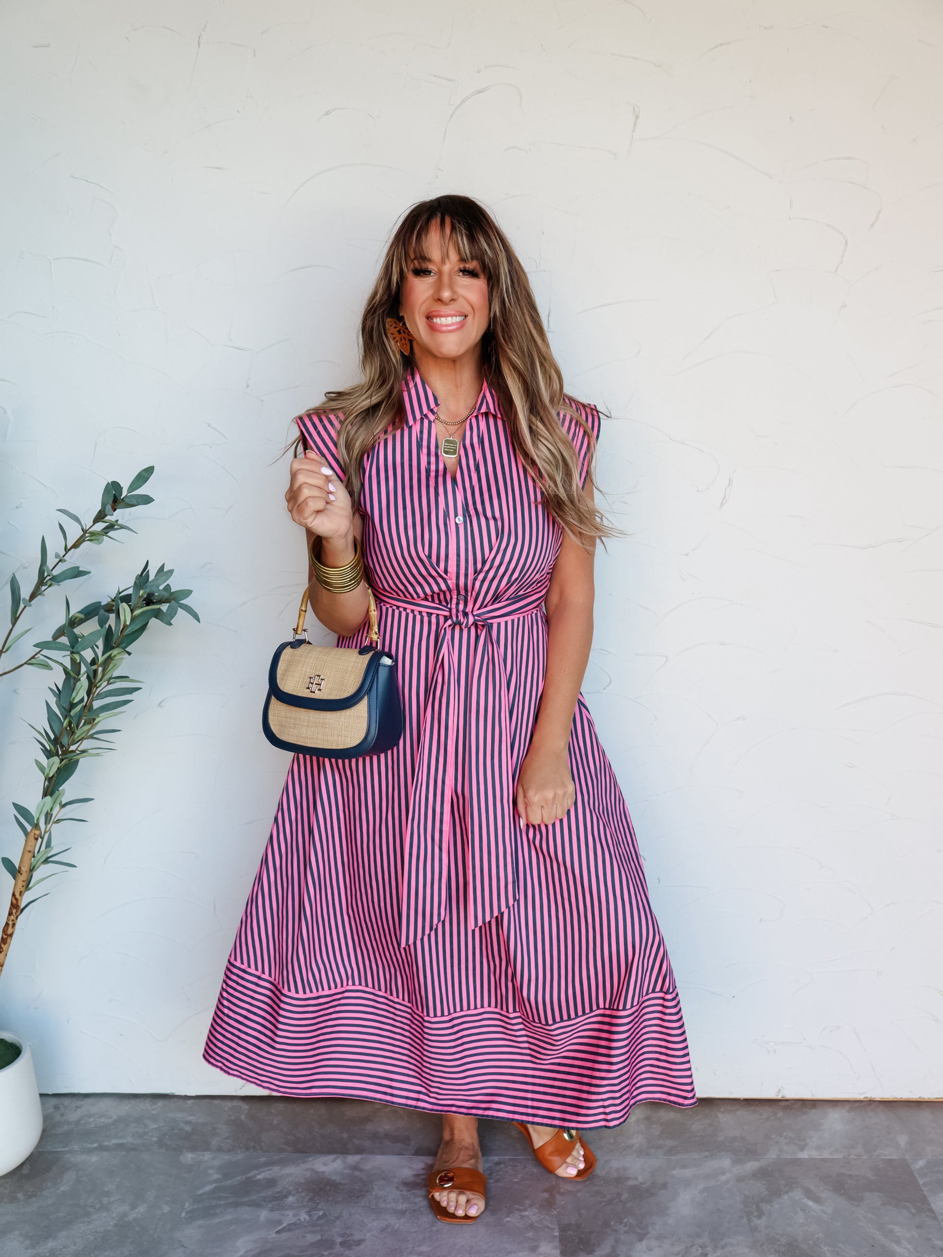 A full-length photograph of a smiling brunette woman with bangs posing against a textured white wall. She is wearing a button-up dark pink and navy blue vertically striped midi shirt dress made from a 100% cotton fabric, featuring a collar, cap sleeves, a matching tie belt at the waist, and horizontal stripes at the hemline, styled with brown slides and a navy and straw handbag.