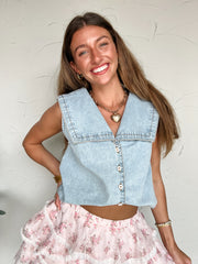 Woman wearing a light blue denim top over a floral skirt against a white wall.