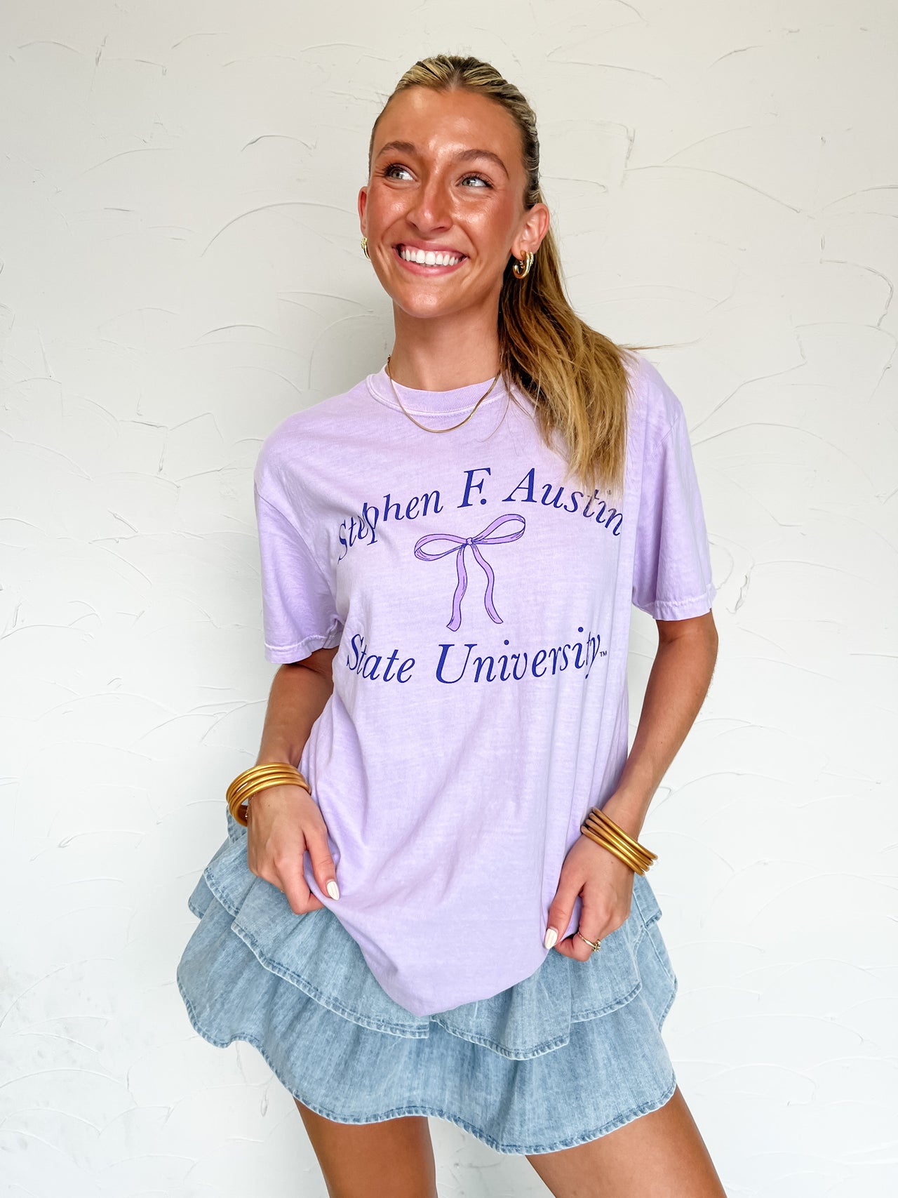 Woman wearing a lavender 'Stephen F. Austin State University' with a bow accent t-shirt and denim skirt against a white background