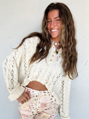 Woman wearing a cream lace eyelet sweater over a white top with floral pants against a white background