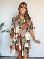 A portrait photograph of a smiling woman with long blonde hair with bangs posing against a textured off-white wall next to a potted olive plant with one hand in her pocket. She is wearing a short-sleeved 100% cotton mini dress with a vibrant olive green, rust, cream, and taupe floral print, featuring a contrast olive collar, a split V-neckline with piping trim, smocked puff sleeves, side pockets, and a tiered drop-waist skirt, styled with a layered pearl necklace, a gold heart pendant, stacked gold bangles,