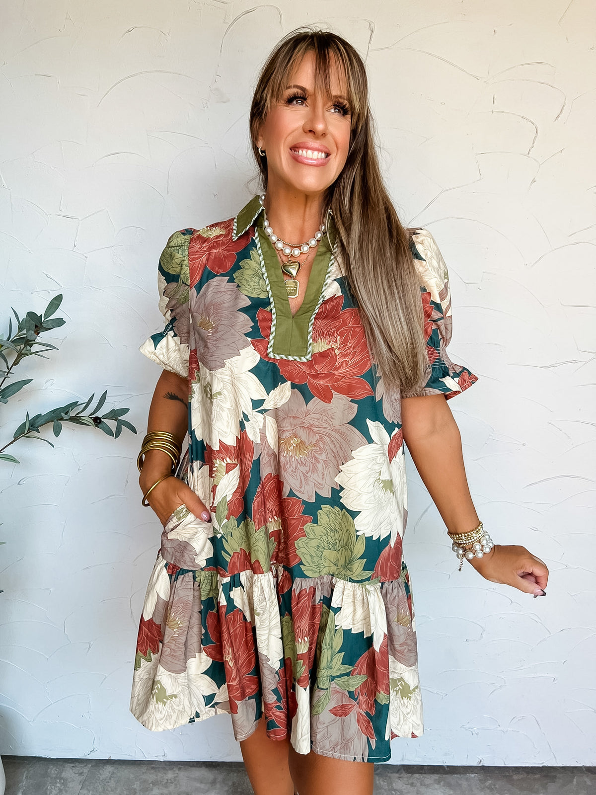 A portrait photograph of a smiling woman with long blonde hair with bangs posing against a textured off-white wall next to a potted olive plant with one hand in her pocket. She is wearing a short-sleeved 100% cotton mini dress with a vibrant olive green, rust, cream, and taupe floral print, featuring a contrast olive collar, a split V-neckline with piping trim, smocked puff sleeves, side pockets, and a tiered drop-waist skirt, styled with a layered pearl necklace, a gold heart pendant, stacked gold bangles,