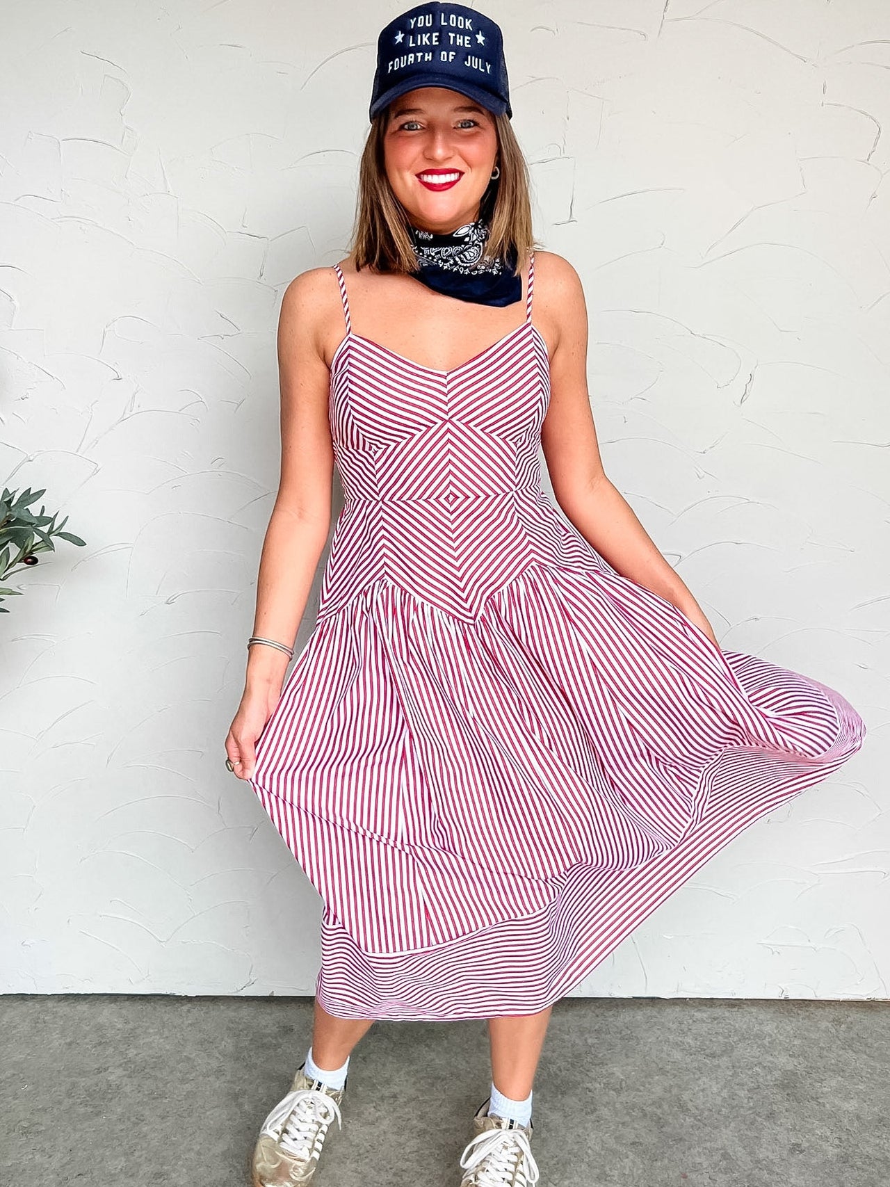 Woman wearing a red and white striped midi dress with a blue cap against a white wall.