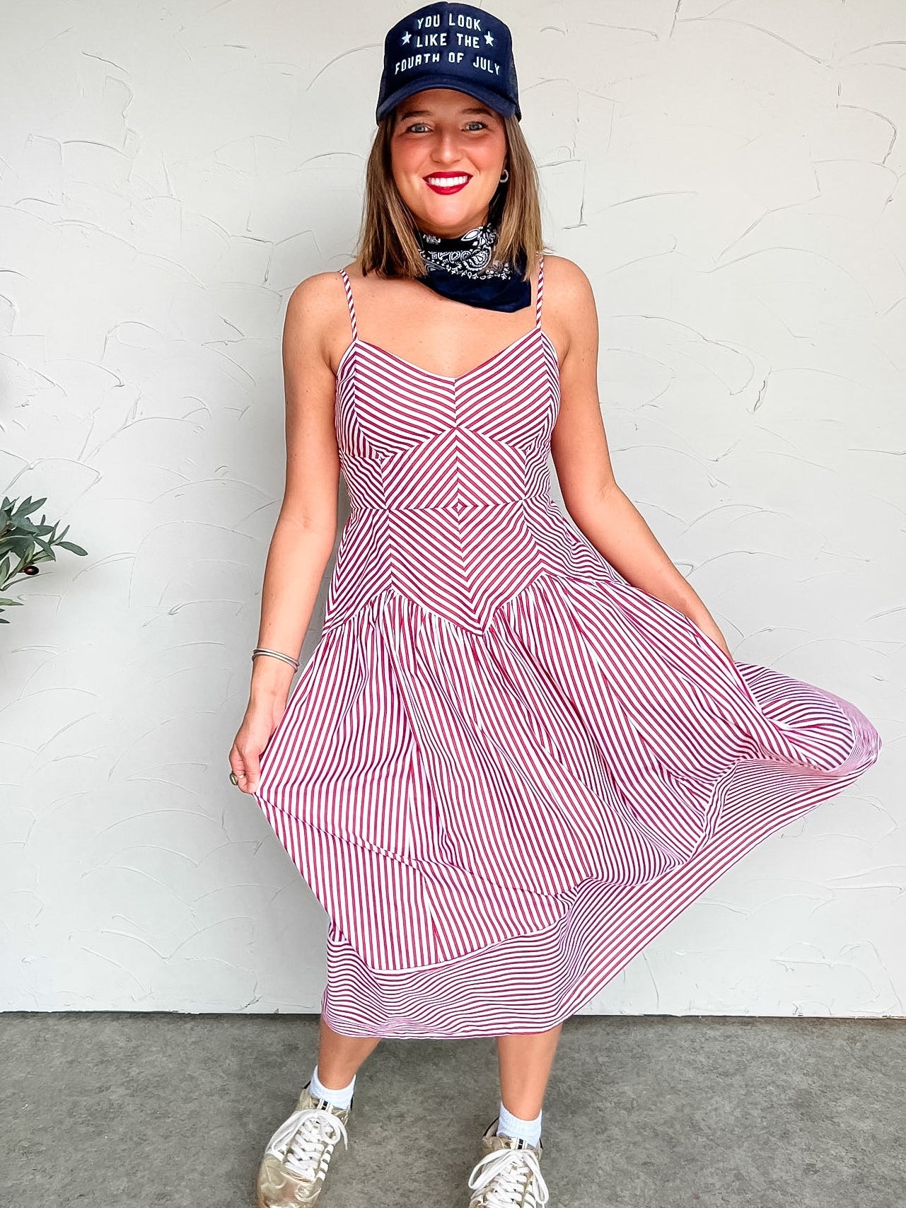Woman wearing a red and white striped midi dress with a blue cap against a white wall.