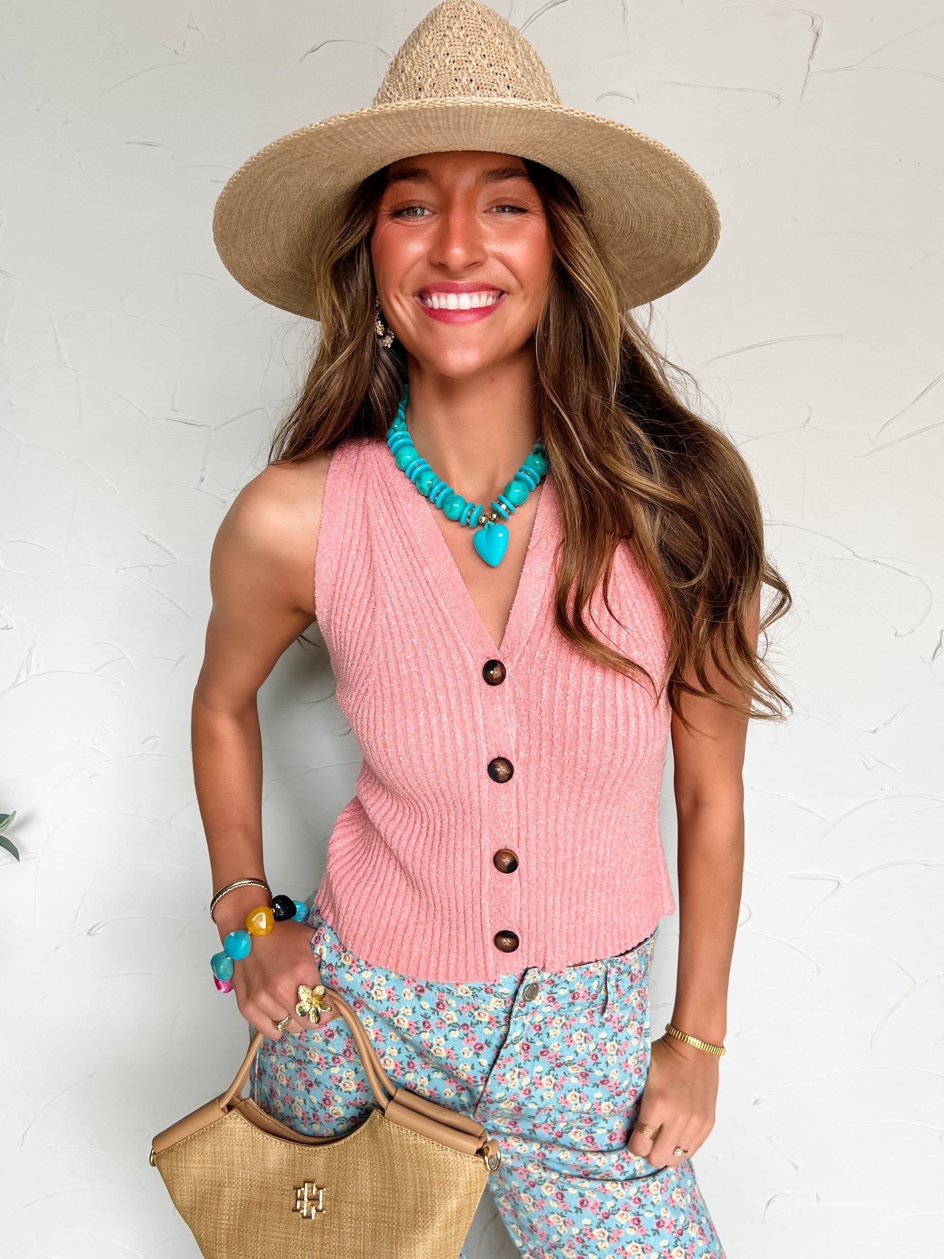 Woman wearing a pink sleeveless sweat vest top, floral pants, straw hat, and carrying a beige handbag against a white background.