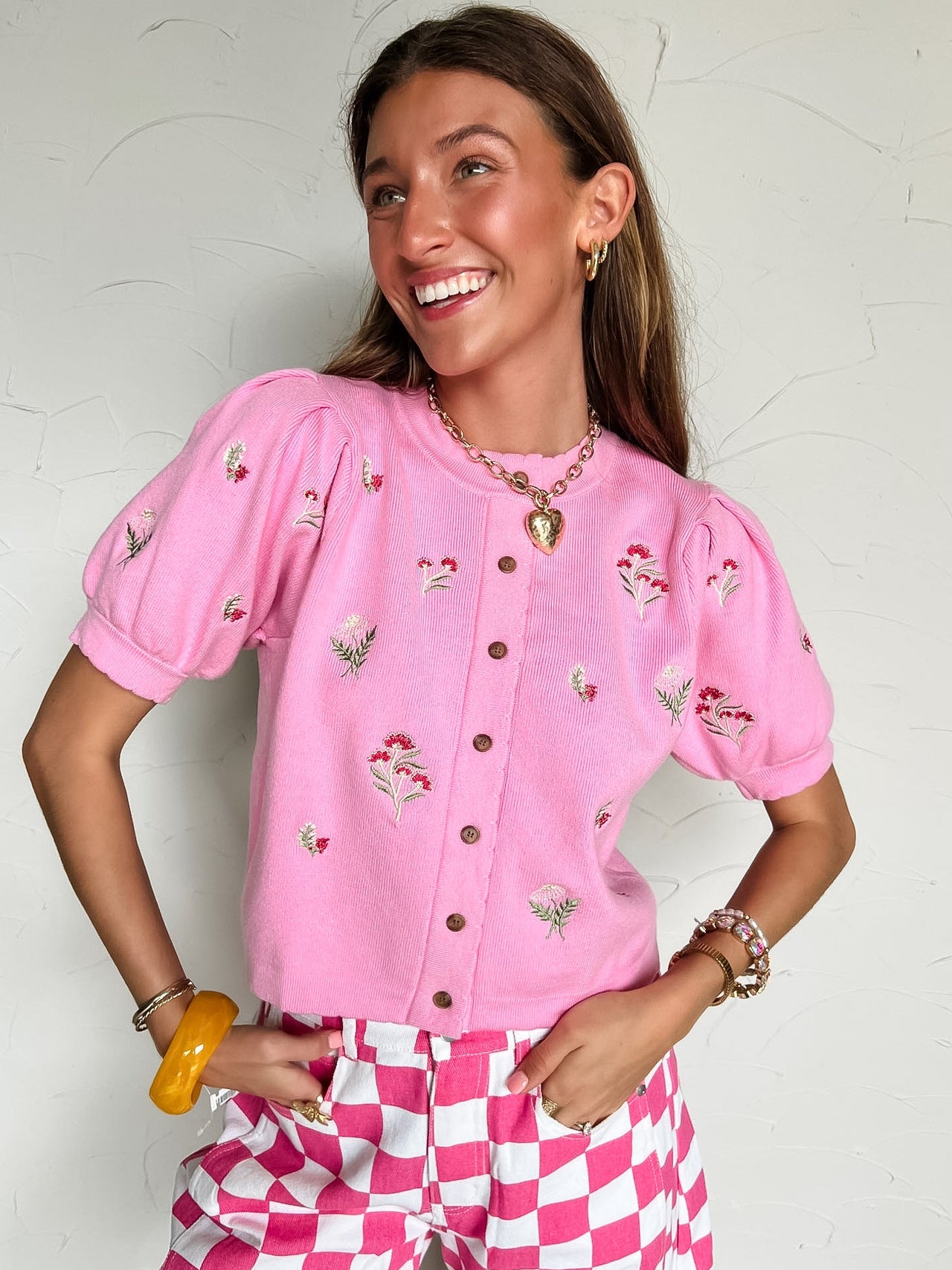 Woman wearing a pink embroidered cardigan and pink and white checkered pants against a white wall.