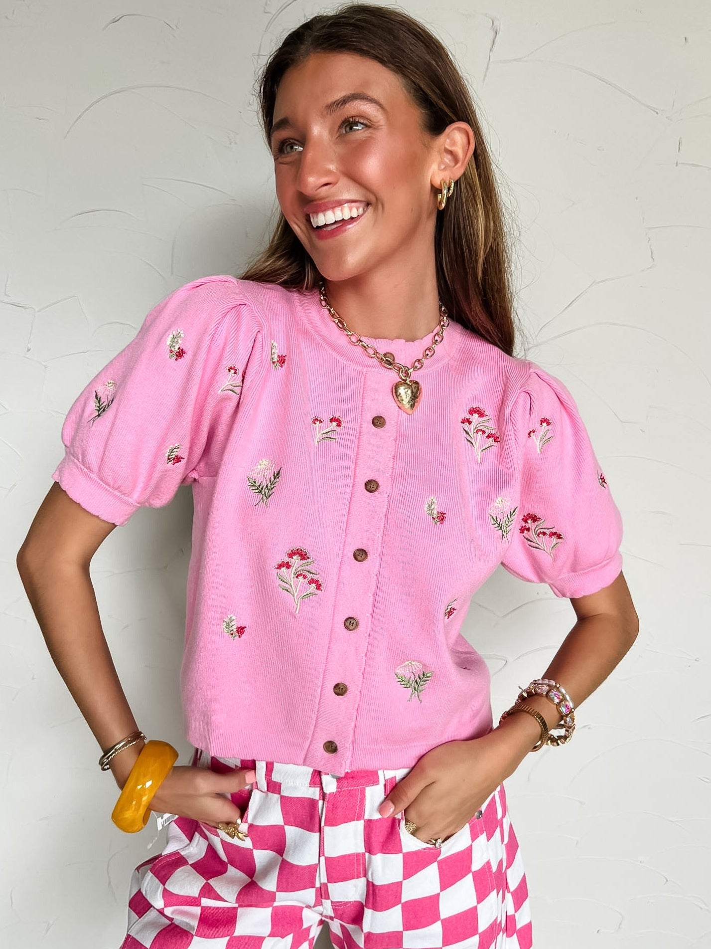 Woman wearing a pink embroidered cardigan and pink and white checkered pants against a white wall.