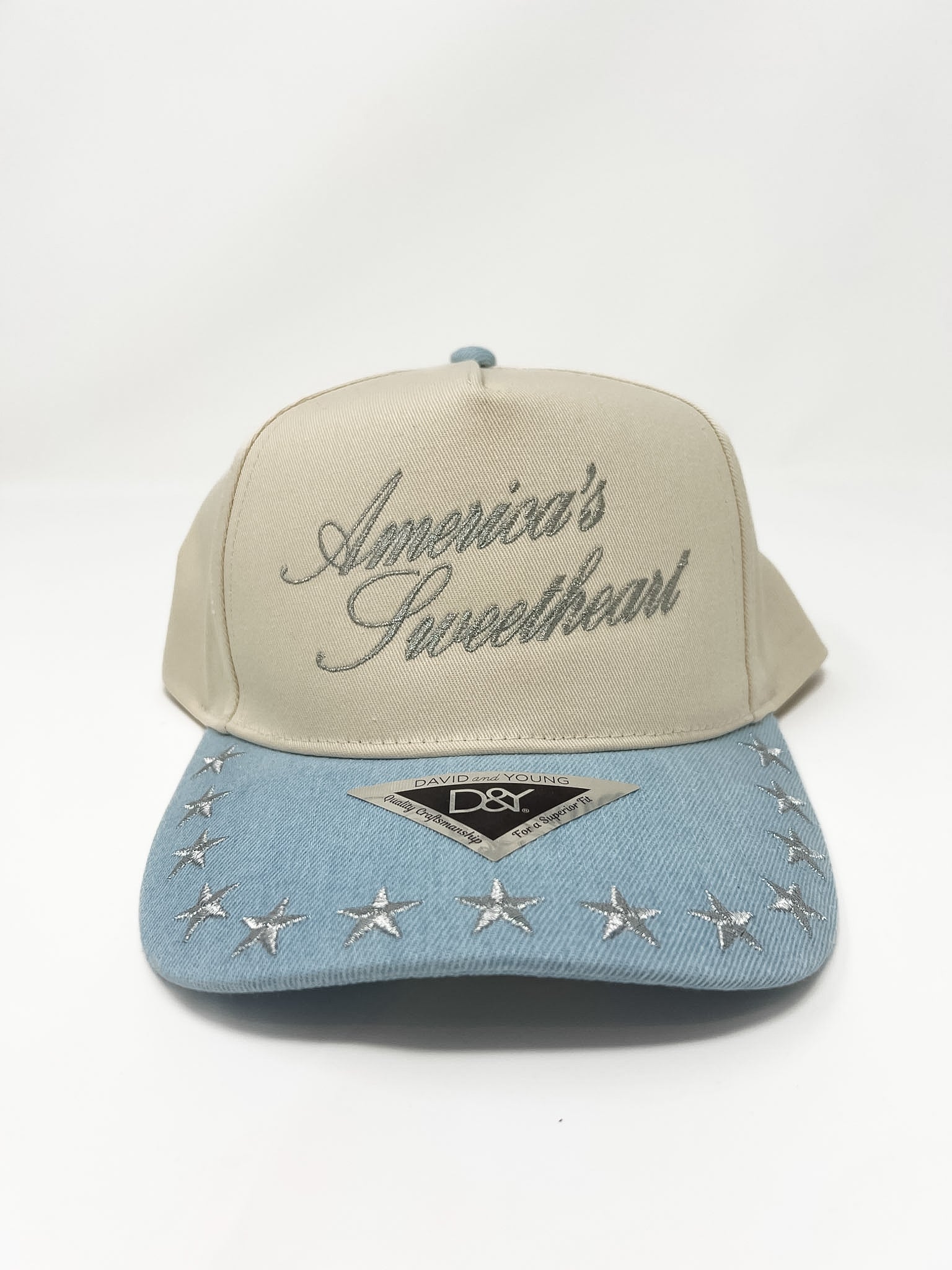 A cream hat that says "America's Sweetheart" with silver script and stitched stars with a denim blue bill.
