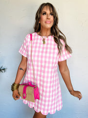 Woman wearing a pink gingham dress with a matching handbag against a light blue wall.