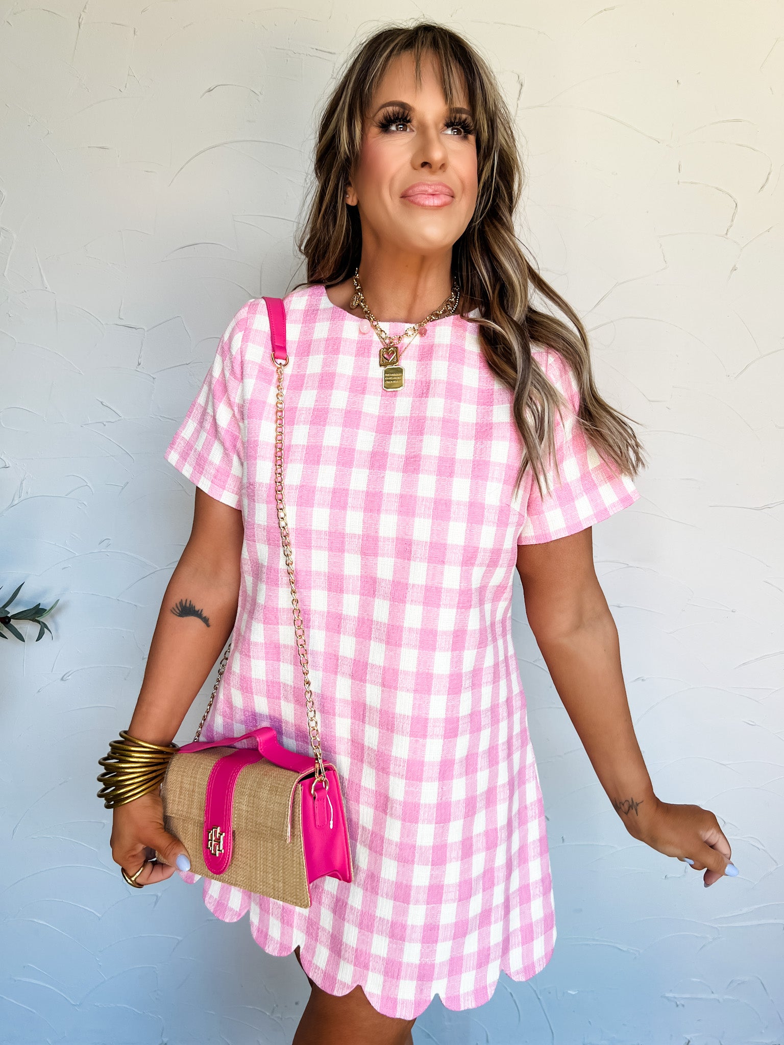 Woman wearing a pink gingham dress with a matching handbag against a light blue wall.