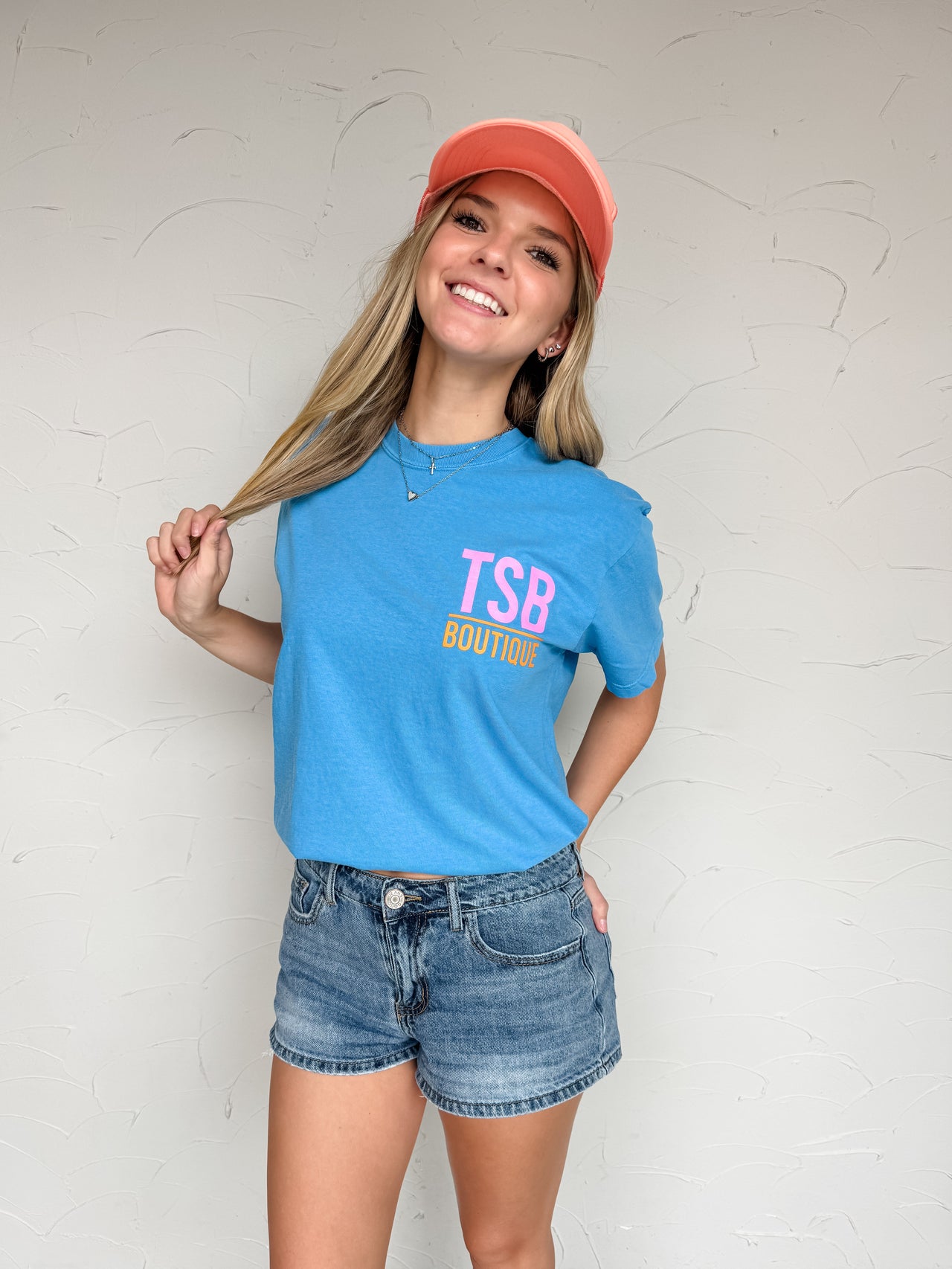 Person wearing a blue t-shirt with 'TSB Boutique' text, denim shorts, and a pink cap against a white wall.